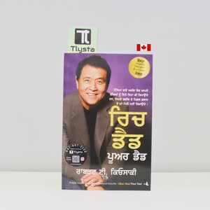 May include: A book titled "Rich Dad Poor Dad" in Punjabi, featuring a portrait of a smiling man on the cover. The book has a purple background with text and a gold seal. A Canadian flag is visible.
