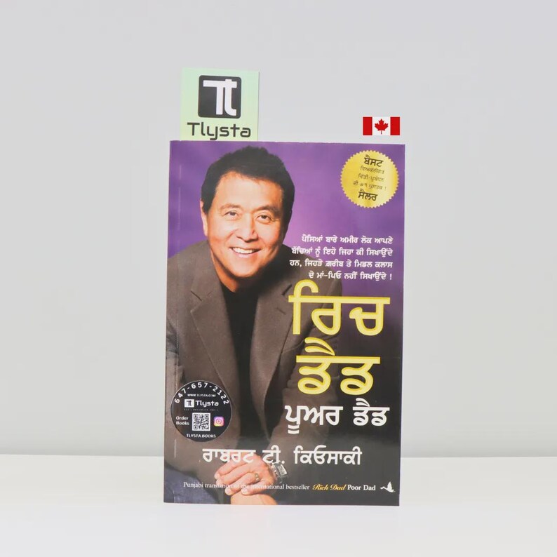 May include: A book titled "Rich Dad Poor Dad" in Punjabi, featuring a portrait of a smiling man on the cover. The book has a purple background with text and a gold seal. A Canadian flag is visible.