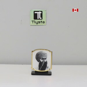 May include: A framed portrait with a black and white image of a man wearing a turban and a beard. The frame has a gold border and sits on a black base. The image is displayed on a white background.