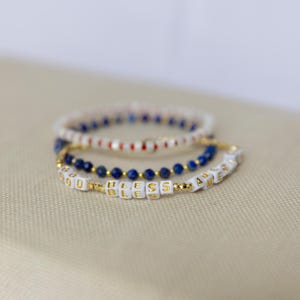May include: Three beaded bracelets. One bracelet has white beads with red and blue accents. Another has dark blue beads. The third bracelet has white letter beads spelling "GOD BLESS YOU" with gold accents.
