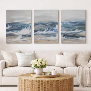 Puede incluir: Tres lienzos que representan olas oceánicas en tonos azules, blancos y beige. Cuelgan sobre un sofá blanco con cojines. Una mesa de centro redonda tejida y una planta en maceta completan el salón. Las obras miden unos 76 cm de alto.