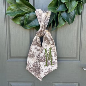 May include: A decorative door hanging featuring a cream-colored fabric with a brown toile pattern, knotted and adorned with a green embroidered "M". The hanging is suspended from a green magnolia leaf wreath, creating a welcoming aesthetic.