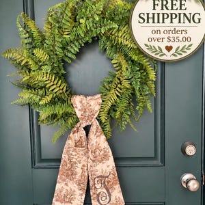 May include: A circular green fern wreath featuring a long, hanging fabric ribbon in a light beige color with a brown toile-style pattern and a large embroidered letter T monogram. The wreath is mounted on a dark green door with metal hardware.