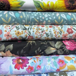 May include: A stack of floral fabric rolls. The top roll features large yellow sunflowers on a white background. Other rolls display various floral patterns in colors like teal, peach, black, and purple. The fabrics are likely cotton or a similar material.