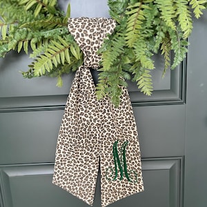 May include: A fabric ribbon featuring a brown and black leopard print pattern with a dark green embroidered monogram letter M. The ribbon is tied in a knot and hangs down with two long tails, positioned over a green fern wreath.