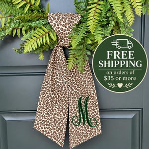 May include: A decorative wreath featuring green fern-like foliage and a long, hanging fabric ribbon with a brown and black leopard print pattern. The ribbon is embroidered with a large, dark green cursive letter M monogram on one side.