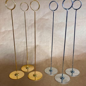 Könnte beinhalten: Eine Auswahl an Tischkartenhaltern aus Metall. Sie haben einen kreisförmigen Sockel, einen schlanken Stiel und einen Ring oben. Einige sind goldfarben, andere silberfarben. Die Halter sind vor einem neutralen Hintergrund angeordnet.