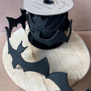 Op de afbeelding: Een rol zwart viltlint met vleermuisuitsparingen. Het lint is gemarkeerd met "d. stevens fine ribbons". De vleermuizen zijn gelijkmatig verdeeld, wat een Halloween-thema creëert. De rol staat op een houten oppervlak.