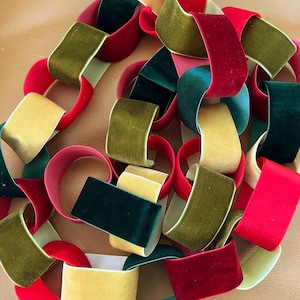May include: A decorative chain constructed from interlocking loops of velvet ribbon in shades of red, dark green, olive green, and gold. The individual links are formed by folding the fabric strips into circles, creating a continuous, flexible garland.