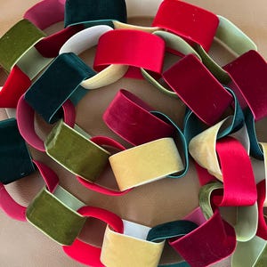 Könnte beinhalten: Eine dekorative Kette aus ineinandergreifenden Samtschlaufen in verschiedenen Farben, darunter leuchtendes Rot, Dunkelrot, Waldgrün, Olivgrün und Creme. Das Samtmaterial hat eine weiche, lichtreflektierende Textur und bildet eine flexible Girlande.