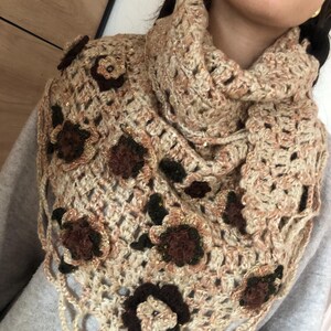 May include: A beige and brown crocheted cowl scarf with floral embellishments. The scarf features a triangular shape with a scalloped edge and is adorned with dark brown and beige flowers. The yarn has a subtle sparkle.