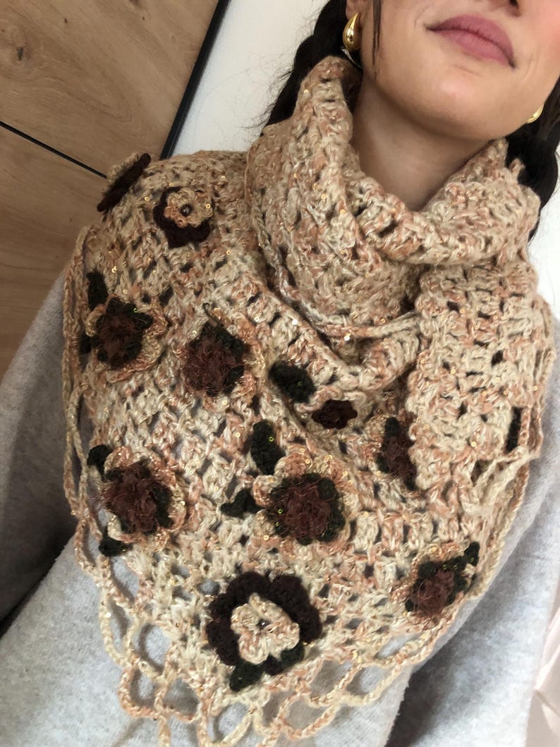 May include: A beige and brown crocheted cowl scarf with floral embellishments. The scarf features a triangular shape with a scalloped edge and is adorned with dark brown and beige flowers. The yarn has a subtle sparkle.