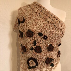 May include: A beige and brown crochet shawl with a floral design. The shawl is draped over a mannequin, showcasing its intricate openwork and decorative dark brown flowers with sequin accents. The edges have a scalloped trim.