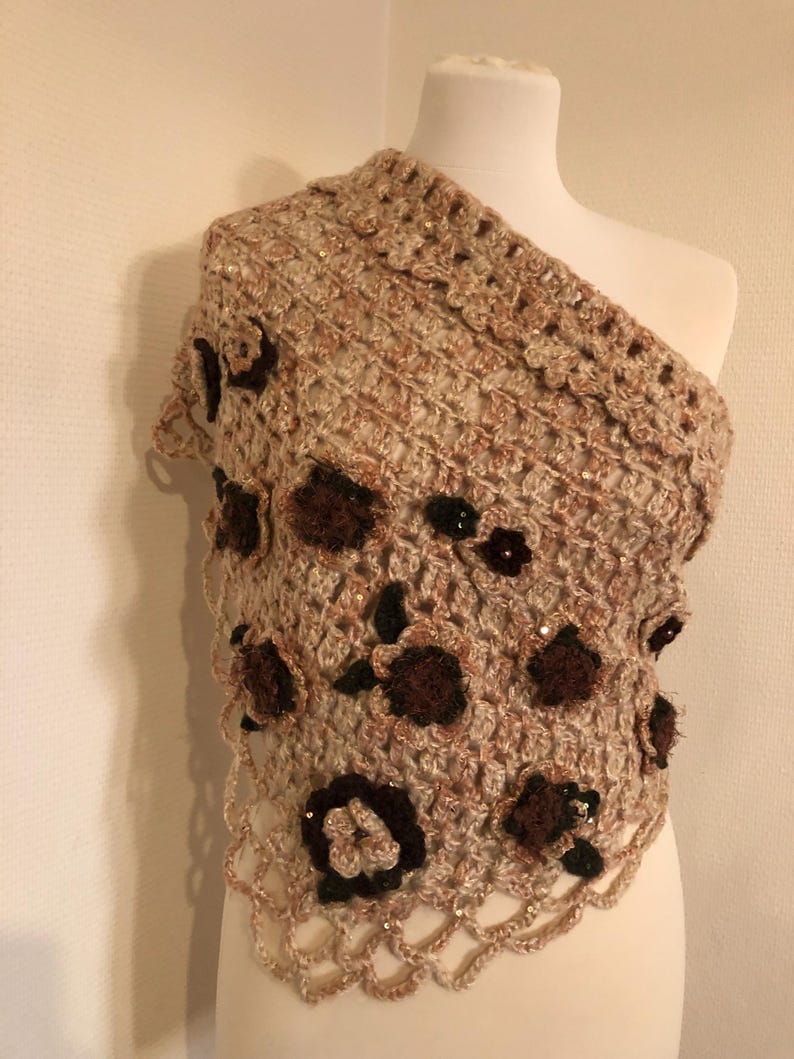 May include: A beige and brown crochet shawl with a floral design. The shawl is draped over a mannequin, showcasing its intricate openwork and decorative dark brown flowers with sequin accents. The edges have a scalloped trim.
