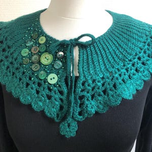 May include: A teal crocheted collar with a scalloped edge and a tie closure. The collar is decorated with various green buttons and small beads. The collar is displayed on a black top.