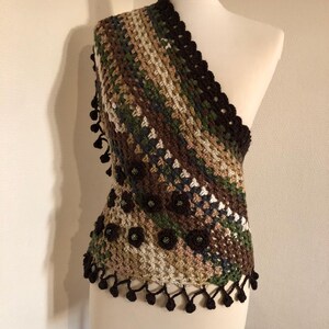 May include: A handmade crochet shawl with a diagonal striped design. The colour scheme features brown, green, and beige tones. The shawl is embellished with dark brown floral accents and a decorative fringe. The garment is displayed on a mannequin.