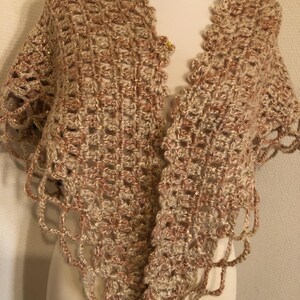May include: A crocheted shawl in shades of beige and light brown, with a scalloped edge. The open weave design features a subtle sparkle. The shawl is draped over a mannequin, showcasing its shape and texture.