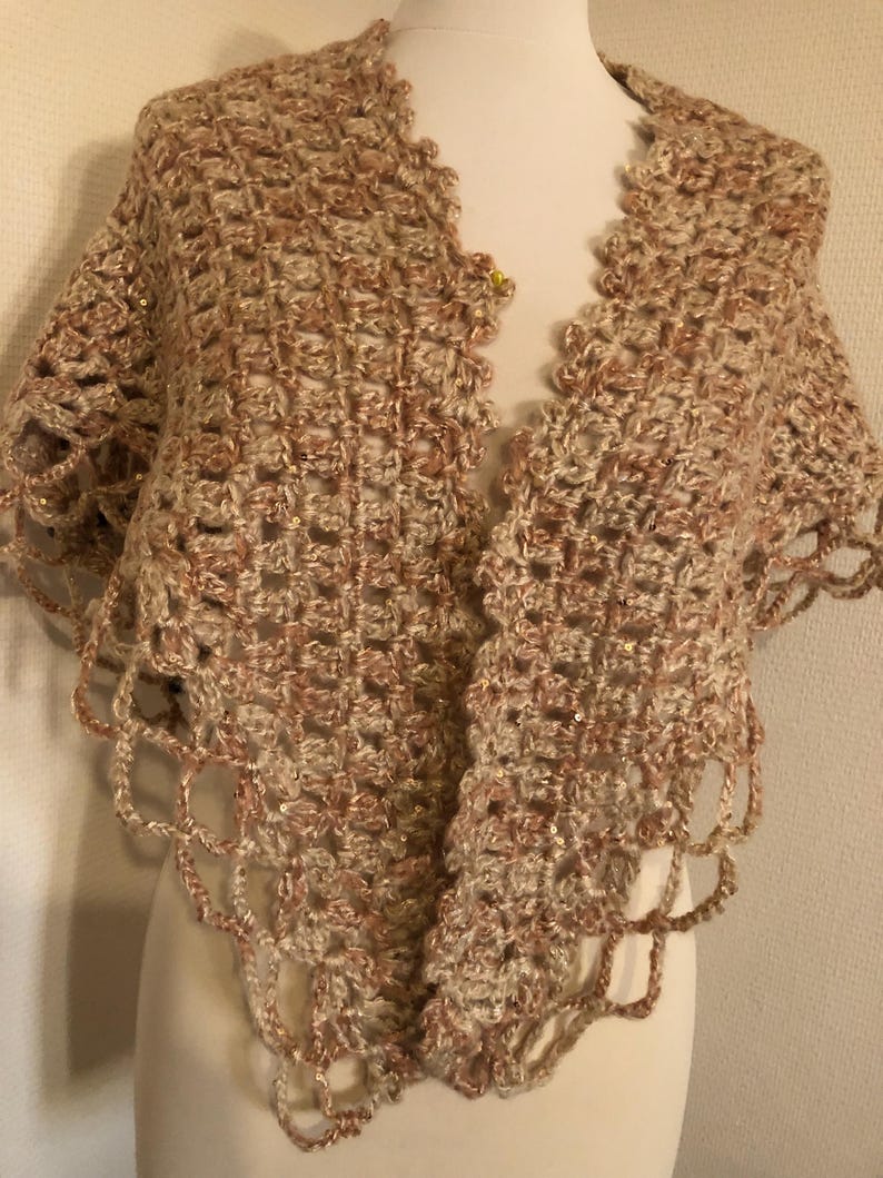 May include: A crocheted shawl in shades of beige and light brown, with a scalloped edge. The open weave design features a subtle sparkle. The shawl is draped over a mannequin, showcasing its shape and texture.