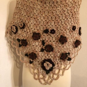 May include: A handmade crochet shawl in shades of beige and brown, adorned with dark brown floral embellishments. The shawl features a scalloped edge and is displayed on a mannequin, showcasing its intricate openwork design.