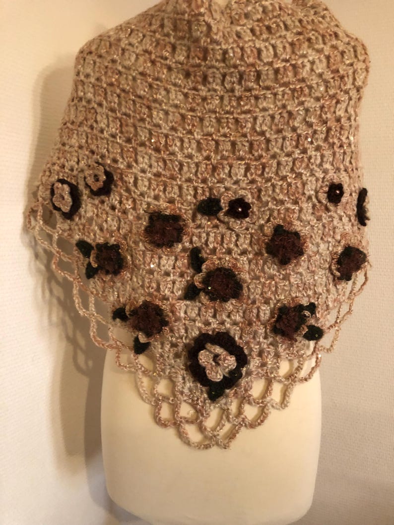 May include: A handmade crochet shawl in shades of beige and brown, adorned with dark brown floral embellishments. The shawl features a scalloped edge and is displayed on a mannequin, showcasing its intricate openwork design.