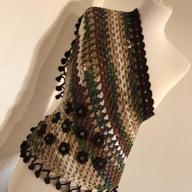 May include: A handmade crochet vest in a camouflage pattern of brown, green, and beige. The vest has a scalloped black trim and is decorated with small, dark brown flower appliqu&eacute;s. Black tassels hang from the bottom edge, adding a decorative touch.