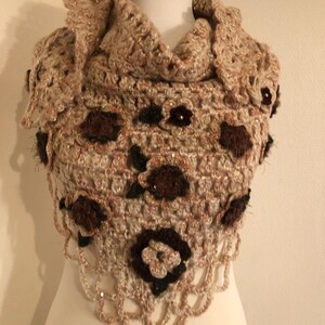 May include: A handmade, beige crochet shawl with brown floral accents and a scalloped edge. The shawl is displayed on a mannequin and features a textured design with a touch of sparkle, showcasing a cozy, bohemian style.