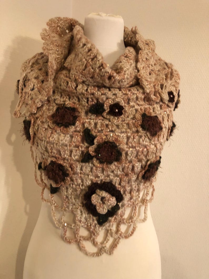May include: A handmade, beige crochet shawl with brown floral accents and a scalloped edge. The shawl is displayed on a mannequin and features a textured design with a touch of sparkle, showcasing a cozy, bohemian style.