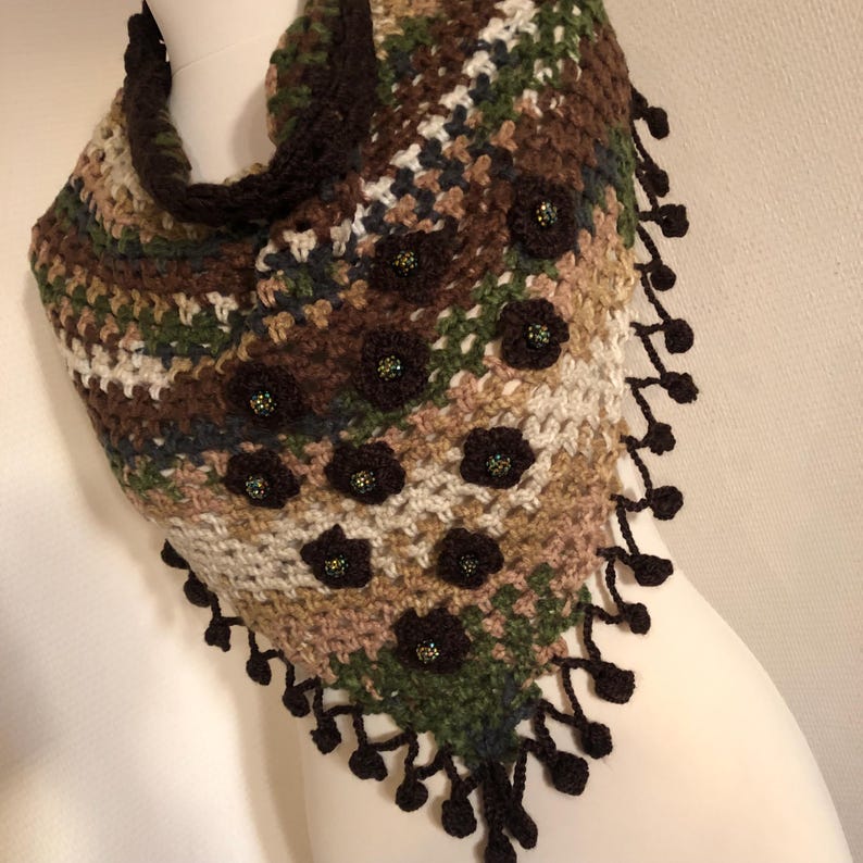 May include: A handmade crochet shawl in a triangular shape, featuring a mix of brown, green, and cream colours. The shawl is adorned with dark brown flower appliqu&eacute;s and a decorative fringe along the edges. The shawl is displayed on a mannequin.