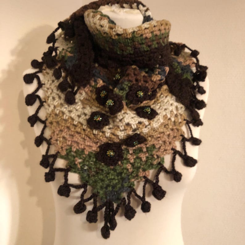 May include: A handcrafted crochet shawl in brown, beige, and green hues, decorated with dark brown flower appliqu&eacute;s and beaded details. The shawl has a fringed edge with small, dark brown bobbles. The item is displayed on a mannequin.