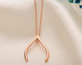 May include: Rose gold wishbone pendant necklace. The necklace features a delicate chain and a pendant shaped like a wishbone. The jewelry is displayed on a satin surface with a glass container and blue flowers in the background.