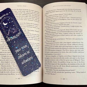 May include: A rectangular bookmark with a dark blue night sky design featuring white stars, a crescent moon, and mountain silhouettes. The text reads BOUND BY STARLIGHT and MAY YOUR DREAMS BE ANSWERED. The bookmark is placed inside an open paperback book.