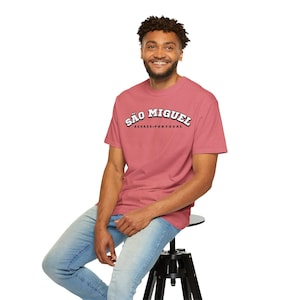 Puede incluir: Una persona con una camiseta rosa polvoriento con el texto "SÃO MIGUEL AÇORES-PORTUGAL" en un diseño blanco y negro. La persona está sentada en un taburete negro y lleva vaqueros azules.
