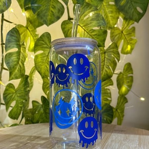 Può includere: Un bicchiere di vetro trasparente a forma di lattina con coperchio in bambù e cannuccia di vetro trasparente. Il bicchiere è decorato con grafiche di faccine sorridenti blu. Lo sfondo presenta foglie verdi e una superficie in legno.