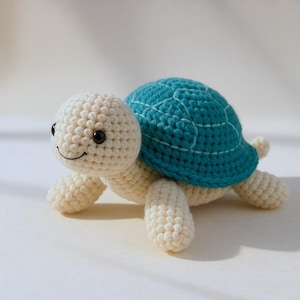 Puede incluir: Una tortuga de crochet hecha a mano, con cuerpo color crema y caparazón turquesa. La tortuga tiene ojos negros y una boca sonriente. El juguete es suave y parece tierno.