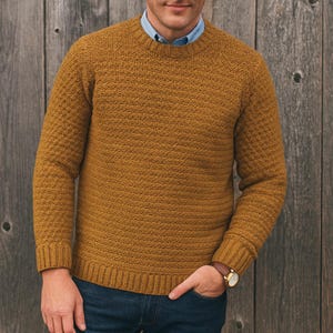 Op de afbeelding: Mosterdgele trui met ronde hals en een textuurgebreid patroon. De trui heeft lange mouwen en geribde manchetten en zoom. De persoon draagt er een lichtblauw overhemd met kraag onder en een blauwe jeans. Een horloge is zichtbaar om de pols.