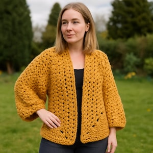 May include: Mustard yellow crochet cardigan with three-quarter sleeves. The open-front sweater has a loose, textured weave. The garment is worn over a black top. The background is a blurred outdoor scene with green foliage.