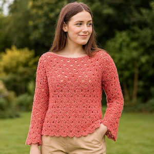 May include: A coral-colored crochet sweater with a boat neck and long sleeves. The sweater has a scalloped hemline and a delicate openwork pattern. The sleeves flare slightly at the wrist. The background is a blurred green outdoor scene.