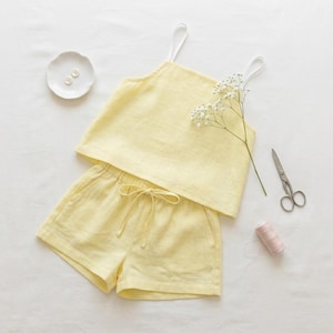Könnte beinhalten: Ein hellgelbes zweiteiliges Set bestehend aus einem ärmellosen Camisole-Top mit dünnen weißen Trägern und einer passenden Shorts mit elastischem Kordelzug. Das Set wird zusammen mit einer kleinen weißen Schale mit zwei Knöpfen und einer Schere gezeigt.