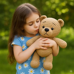 以下が含まれることがあります： 黒い目と黒い鼻を持つベージュのかぎ針編みのテディベアが、デイジー柄の青いシャツを着た人に抱かれています。クマは縫い付けられた口をしており、キスをされています。背景は柔らかな緑色です。