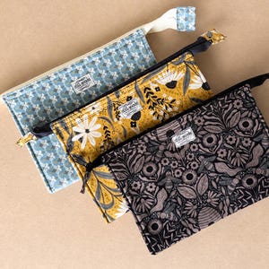 May include: Three rectangular zippered pouches featuring distinct patterns: a blue geometric print, a mustard yellow floral design with white daisies, and a black and gray botanical print with bees. Each pouch has a small white label reading Still Goods.