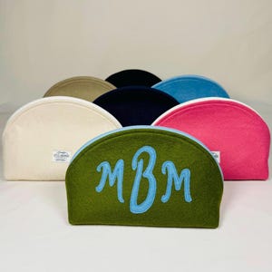 May include: A collection of half-moon shaped cosmetic bags in various colors, including green, pink, blue, and cream. The front bag is olive green with light blue embroidered initials. Each bag has a zipper closure and a small tag.