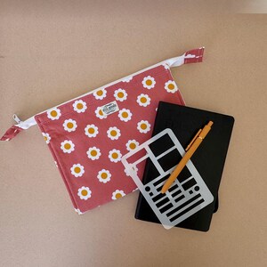 May include: A rectangular zippered pouch in dusty rose fabric with a white and yellow daisy pattern and a small white label reading still-goods. Beside it lies a black notebook, a yellow ballpoint pen, and a white plastic stencil sheet.