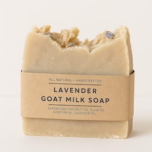 May include: A rectangular bar of lavender goat milk soap with a torn top, wrapped in a brown paper band. The band reads "LAVENDER GOAT MILK SOAP" and "ALL NATURAL + HANDCRAFTED". The soap contains lavender buds.