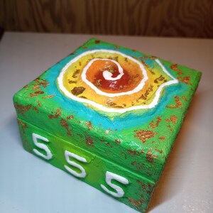 May include: A square wooden box painted in bright green with gold leaf accents. The lid features a raised, textured spiral design in shades of yellow, orange, and red, outlined in white. The front side displays the white number 555 in relief.
