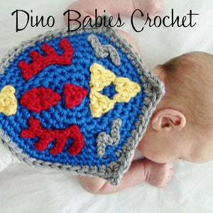 May include: A crocheted baby costume in the shape of a shield with a blue background, red and yellow accents, and a gray border. The shield is worn by a baby lying on its stomach.