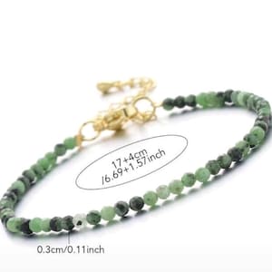 May include: A beaded bracelet featuring small, faceted green and black stones. The bracelet includes a gold-toned metal lobster clasp and an extender chain. Text on the image indicates a length of 17 cm plus 4 cm and a bead size of 0.3 cm.
