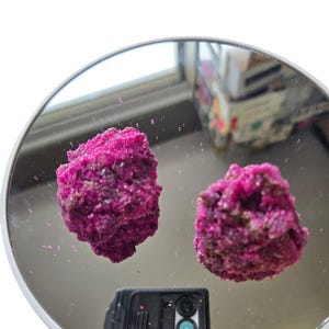 Peut inclure: Deux amas minéraux cristallins magenta vif avec une surface rugueuse et texturée. Ces formations rocheuses irrégulières et dentelées sont placées sur une surface miroir circulaire et réfléchissante, créant un reflet clair des objets.