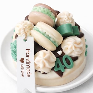 Puede incluir: Un pastel decorativo con glaseado blanco, detalles de chocolate y macarons y remolinos de glaseado verdes y crema. El pastel está decorado con el número 40 y una etiqueta que dice "Handmade with love".