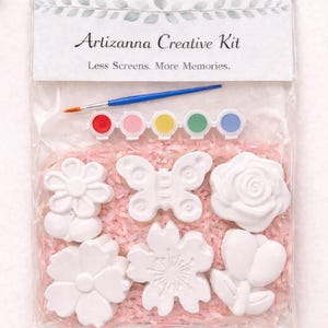 Puede incluir: Una bolsa de plástico transparente contiene un kit creativo con el texto "Artizanna Creative Kit" y "Less Screens. More Memories." En el interior hay formas de flores y mariposas blancas, botes de pintura y un pincel. El texto "Paint your own flowers!" está en la parte inferior.