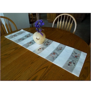May include: A white table runner with a silver and brown floral pattern. The runner is on a wooden table with a wooden vase with purple flowers in the center.